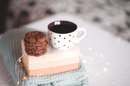 Cup of black tea with chocolate cookies staying on stack of book and knitted sweater closeup in bed....