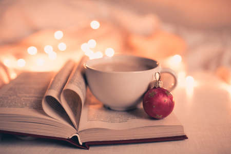 Cup of coffee with open book and heart shape pages over Christmas lights close up. Winter holidays.