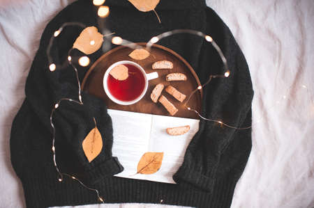 Knitted sweater with wooden tray, open book, cup of tea and cookies closeup over glowing lights. Top view. Good morning. Autumn season.の写真素材