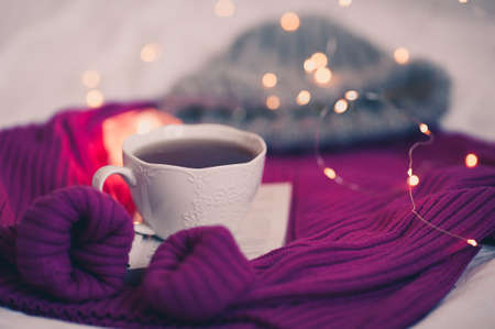 White cup of coffee staying on open book and knitted sweater over Christmas lights close up. Winter season.の写真素材