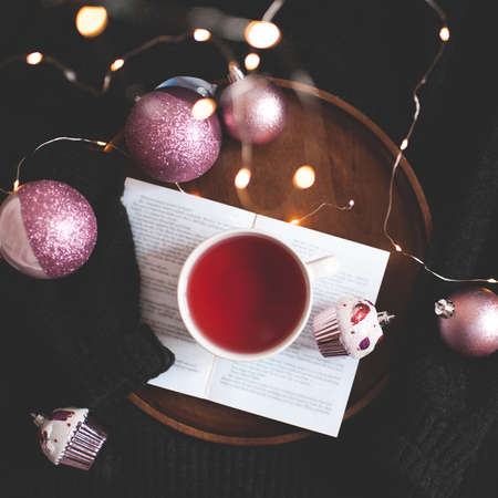 Cup of tasty tea on open book with Christmas decorations over black sweater background close up....