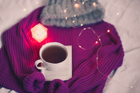 Fresh coffee in white cup staying on open book with candles on knitted sweater in bed closeup. Winter time.の写真素材
