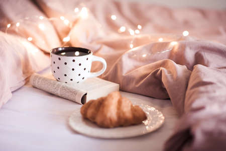 Fresh black tea in mug staying on open book with croissant in bed glowing lights closeup. Winter...