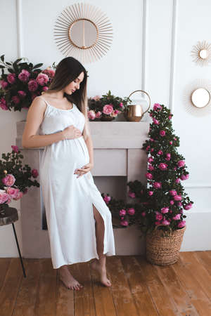Beautiful pregnant woman 20-25 year old wearing elegant white dress posing in room with rose flowers...