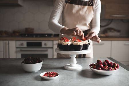 Woman decorate cupcakes with fruits and cream cheese closeup in kitchen. Working at home. Selective focus.の写真素材