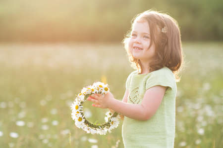 Smiling child girl 2-3 year old making flower wreath posing in field over sunny background closeup. Summer season. Childhood.の写真素材