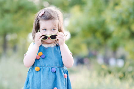 Cute baby girl 2-3 year old posing in park holding flowers closeup. Wearing trendy denim dress....