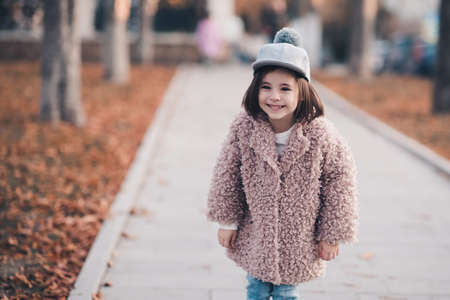 Pretty kid girl 5-6 year old wearing winter coat and stylish hat posing in park outdoors. Autumn season. Childhood. Happiness.の写真素材
