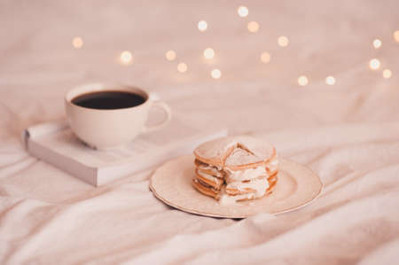Tasty pancakes with cup of coffee and open book over glowing lights in bed closeup.の写真素材