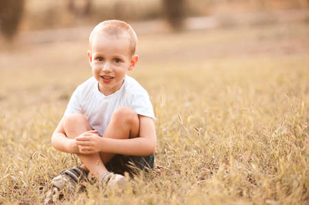 Cute kid boy 3-4 year old wearing casual white t-shirt sitting in meadow outdoors. Childhood.の写真素材
