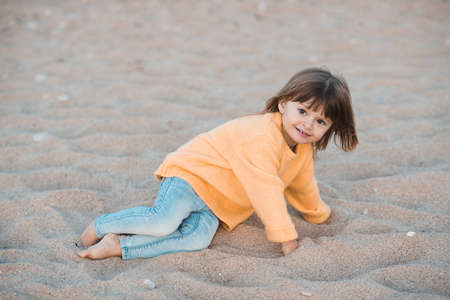 Cute kid girl 2-3 year old wearing knitted yellow sweater and denim pants having fun at beach playing with sand outdoors closeup. Childhood. Autumn season.の写真素材