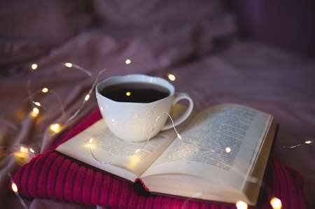 Cup of hot black tea staying on open book and knitted pink sweater over glowing Christmas lights...