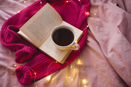 Cup of coffee with open book and knitted clothes over glowing lights in bed close up. Selective focus. Top view.の写真素材
