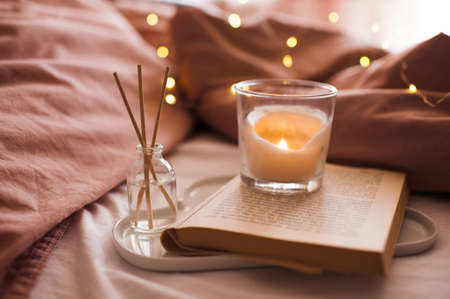 Burning candle with aroma sticks in bottle on tray with open book in bed over glowing Christmas lights close up. Cozy atmosphere at home. Good morning. Selective focus.の写真素材