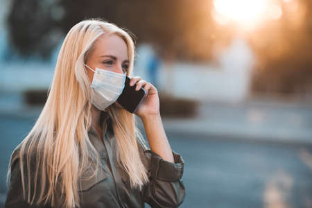 Young blonde woman 24-26 year old wearing medical protective mask talking on phone walking in street outdoors close up. Coronavirus pandemic. Stay at home.の写真素材