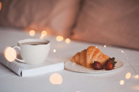 Cup of coffee with open paper book and cake in bed close up over Christmas lights. Good morning.の写真素材