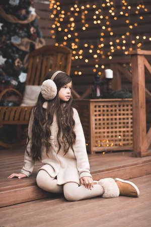 Stylish kid girl 5-6 year old wearing knitted clothes and fluffy headphones sitting outdoors over Christmas lights close up. New Year. Winter holiday season.の写真素材
