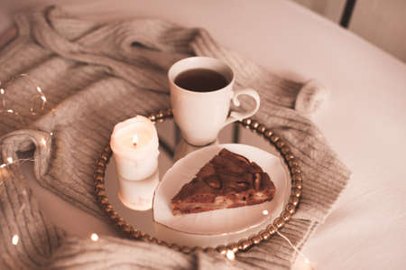 Chocolate pie with cup of tea and burning candle on tray with knitted sweater in bed close up. Good...