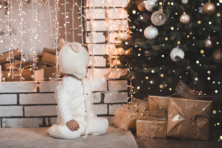 Baby 1-2 year old wearing knitted suit and hat sitting under Christmas tree with glowing lights and decorations close up. Winter holiday season. Childhood.の写真素材