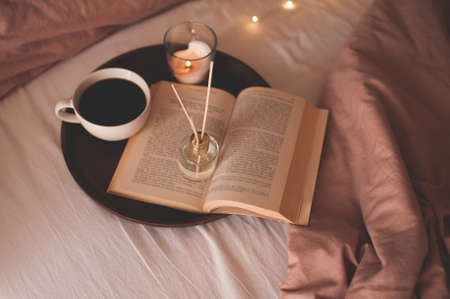 Cup of coffee with open paper book on wooden tray with liquid scented home fragrance in bed over glowing lights close up. Cozy atmosphere concept. Good morning. Freshness.の写真素材