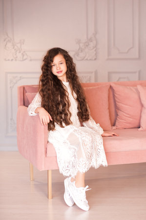 Beautiful child girl 6-7 year old wearing white stylish dress sitting in pink sofa in room closeup. Looking at camera. Childhood. Fashionable.の写真素材