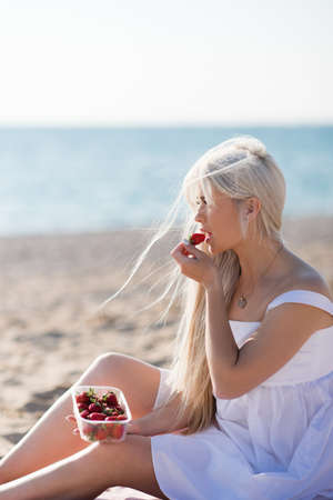Beautiful young woman eating fresh strawberry having picnic over sea shore outdoors. Summer season. Healthy lifestyle.の写真素材