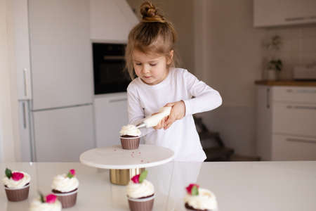 Cute baby girl 3-4 year old making cupcake, decorate with whipped cream on kitchen table at home close up. Childhood.の写真素材