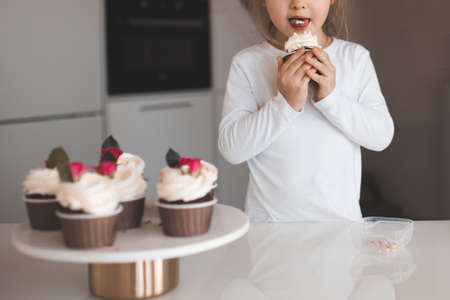 Cute baby girl 2-3 year old eating tasty cupcakes with cream on top at kitchen indoors. Childhood. Selective focus.の写真素材