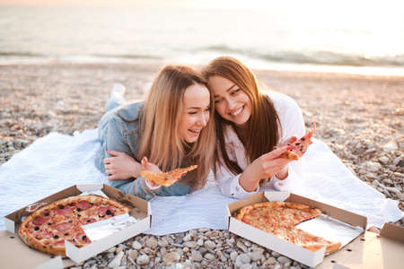 Two young beautiful girlfriends having fun with pizza and wine at beach over sea shore outdoors in sun light. Summer vacation season. Friendship. Happiness. Sisters spend time together at coast line.の写真素材
