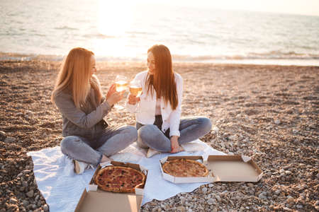 Two young beautiful girlfriends having fun with pizza and wine at beach over sea shore outdoors in sun light. Summer vacation season. Friendship. Happiness. Sisters spend time together at coast line.の写真素材