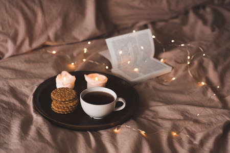 Cup of fresh black tea with stack of biscuits with chocolate filling on tray with burning candles and open paper book in bed over glow lights. Cozy home atmosphere.の写真素材