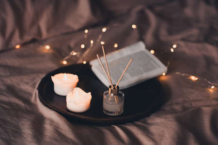 Liquid home fragrance in glass bottle and wood sticks on tray with candles and open paper book in bed close up. Good morning. Relaxation. Selective focus.の写真素材