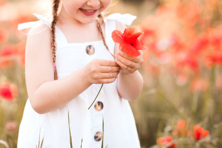 Smiling happy child girl 3-4 year old holding flower stand in poppy meadow outdoors over nature background. Wear white top and straw hat in field. Childhood. Summer season. Happiness. Freedom.の写真素材