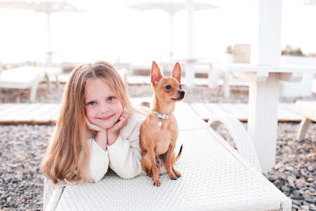Smiling cute blonde kid girl 3-4 year old lying at beach with pet domestic dog outdoors. Childhood. Looking at camera. Heathy lifestyle. Summer vacation season.の写真素材