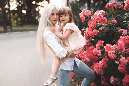 Beautiful adult woman holding baby girl wear stylish dress posing over blooming rose flowers in city street outdoors together. Family concept lifestyle. Motherhood. Parenthood.の写真素材