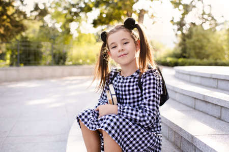 Cute young child girl 5-6 year old wear checkered black and white dress and backpack holding books sitting on stairs outdoors close up. Looking at camera. Back to school. Study concept.の写真素材