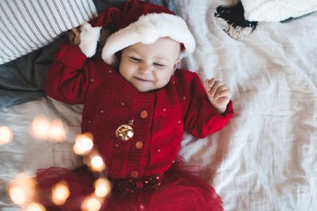 Cute baby girl 1 year old wear red santa claus hat and knit dress lying in bed close up. Smiling little child celebrating christmas holidays. Top view. Childhood. Winter season. Happiness.の写真素材