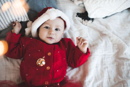 Cute baby girl 1 year old wear red santa claus hat and knit dress lying in bed close up. Smiling...
