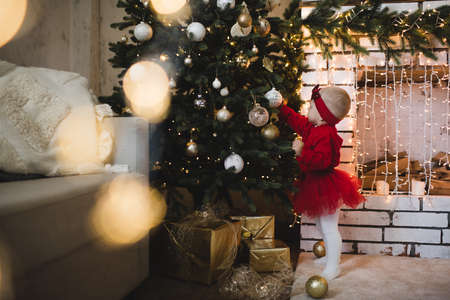 Sweet kid toddler girl playing with xmas tree in room over glow lights indoors. Childhood. Happy New Year. Winter holiday season. Child celebrating. Celebration.の写真素材