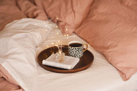Cup of black fresh coffee with liquid aroma perfume in glass bottle with wood sticks and open paper book in tray in bed with pink blacket and duvet. Cozy home atmosphere. Aromatherapy.の写真素材