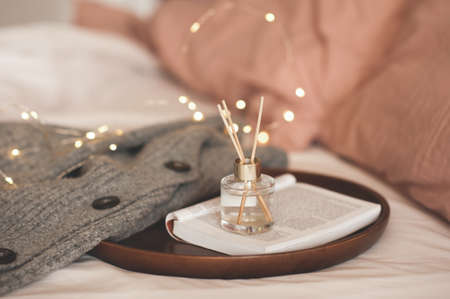 Home liquid perfume in glass bottle in tray with paper open book with knitted testile fabric in bed close up. Winter hygge season. Cozy atmosphere. Aromatherapy.の写真素材