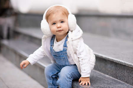 Cute baby girl 1-2 year old wear fluffy white headphones, denim suit pants and knitted sweater posing on city street outdoors. Childhood. Happy little child.の写真素材