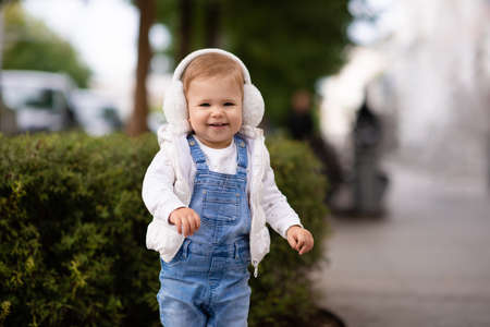 Cute baby girl 1-2 year old wear fluffy white headphones, denim suit pants and knitted sweater posing on city street outdoors. Childhood. Happy little child.の写真素材