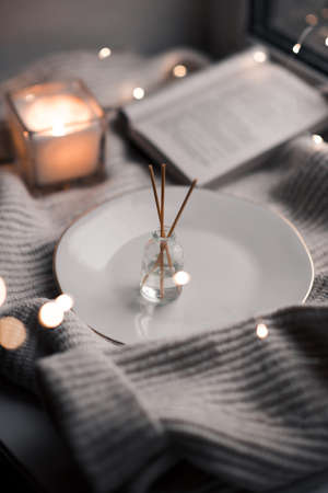 Home liquid perfume in glass bottle and wooden sticks with burning candle and open paper book on knit fabric cloth close up. Cozy warm atmosphere. Winter season.の写真素材