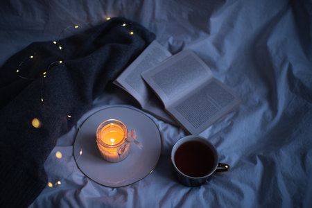 Burning scented candle on ceramic tray with open paper book and knit sweater cloth in bed. Winter cozy hygge atmosphere with cup of black coffee at home. Coziness.の写真素材