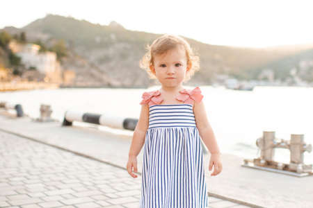Smiling baby girl 1-2 year old wear striped dress and holding walk over wooden pier at sea shore outdoors. Summer season. Childhood. Looking at camera.の写真素材