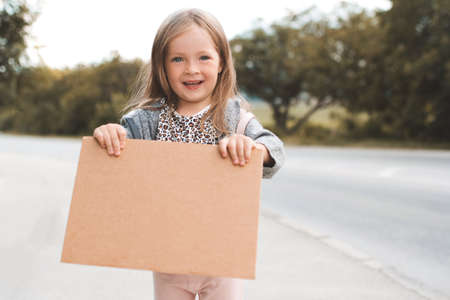 Happy smiling child girl 3-4 year old hold paper craft Paris handwritten sign hitch hiking on road outdoors. Happiness. Little kid toddler travel wear casual clothes and backpack. Summer vacation timeの写真素材