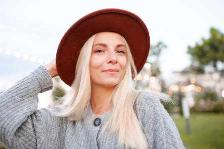 Beautiful blonde smiling adult girl 20-24 year old wear casual warm clothes and hat posing in city park. Autumn season. Romantic lady outdoors. Looking at camera.の写真素材