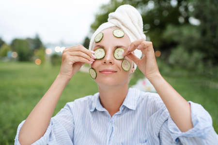 Young adult girl 22-24 year old making facial mask with fresh sliced cucumber wear towel on head...