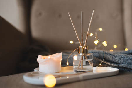 Scented candle and liquid home perfume in glass bottle with wooden sticks staying on tray over...